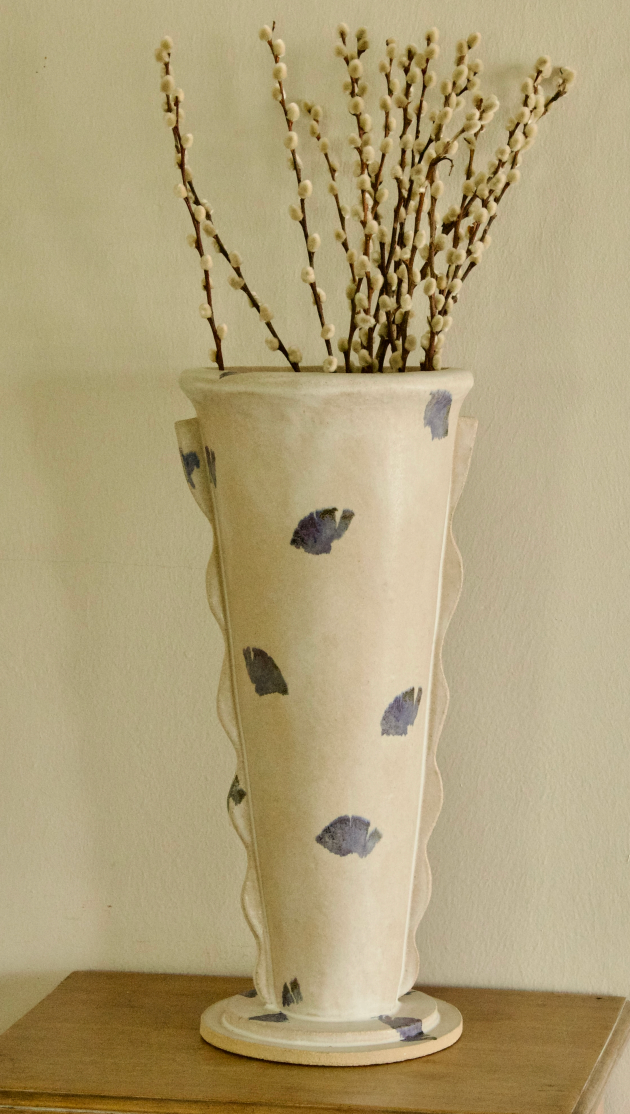 A tall cream-colored ceramic vase with a scalloped rim, decorated with hand-painted blue leaf motifs, filled with dried willow branches and displayed on a wooden surface against a neutral beige wall.
