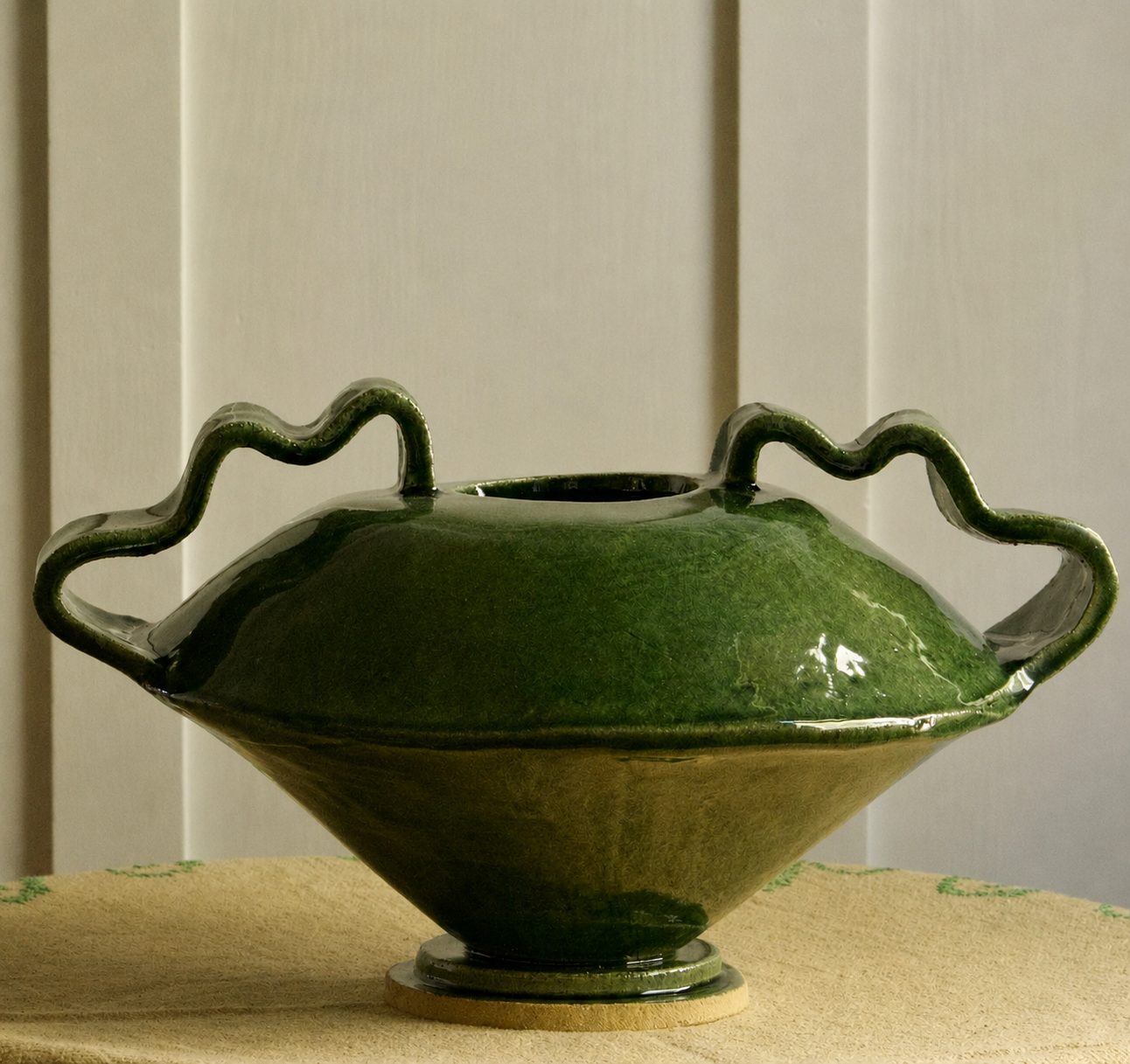 A glossy dark green ceramic vase with a wide, flared base, a small opening at the top, and two wavy loop handles, displayed on a textured beige surface against a light paneled background.