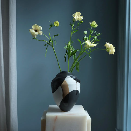 Black and white vase with flowers on a white pedestal against a dark blue wall.