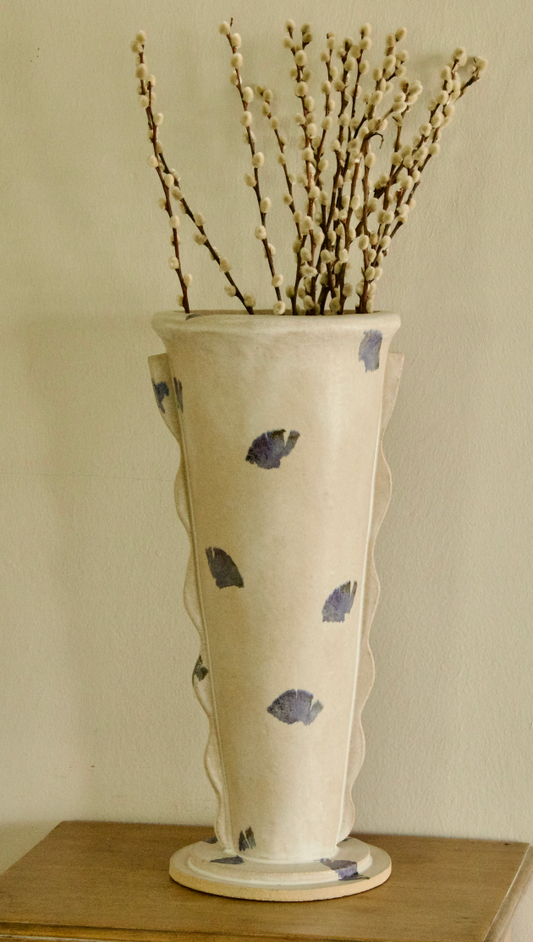 A tall cream-colored ceramic vase with a scalloped rim, decorated with hand-painted blue leaf motifs, filled with dried willow branches and displayed on a wooden surface against a neutral beige wall.